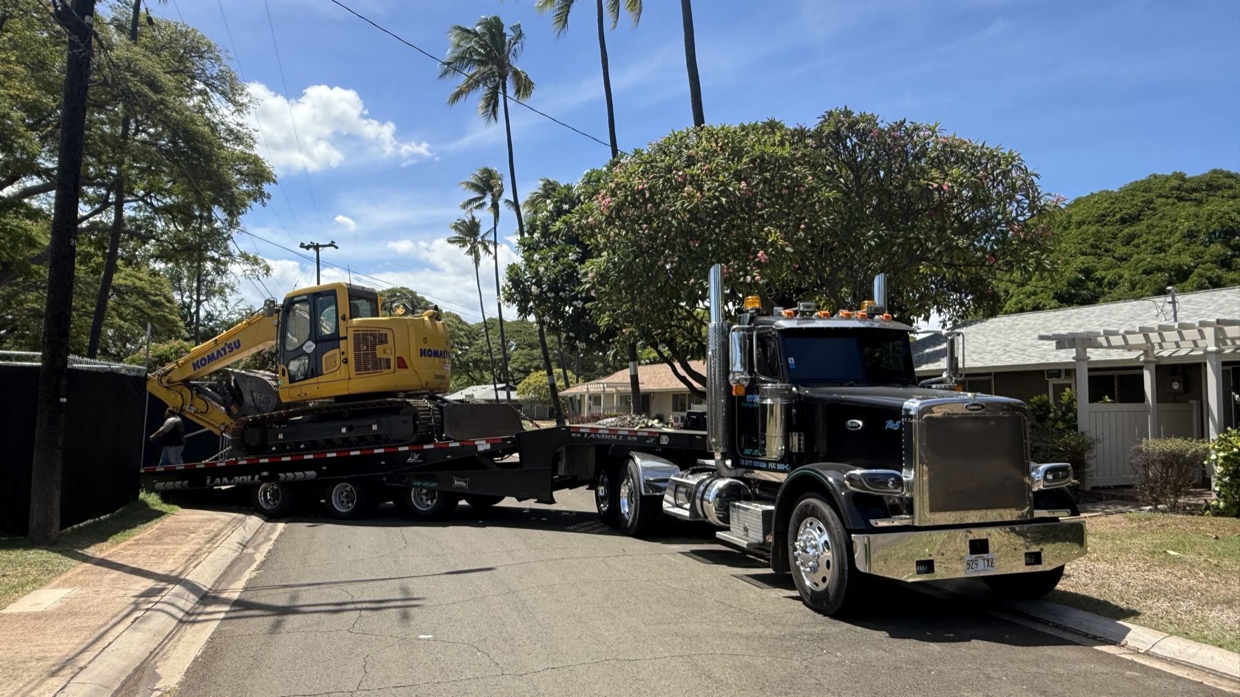 Stone's flatbed transporting a Komatsu excavator through a residential Oahu street